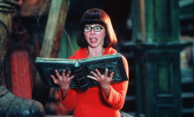 A woman in an orange sweater and black glasses holds a book and screams.
