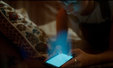 A young adult looks over their phone, which is emitting a strong light toward them. 