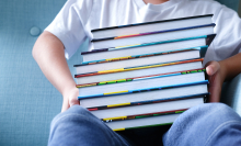 A child sits on a light blue couch holding a large stack of books. 