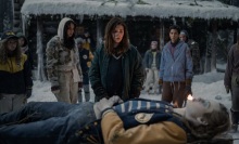A group of young women in winter clothes stare at the corpse of a young woman in a letterman jacket that they've laid out on a pyre.