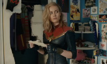Captain Marvel stands in a teen bedroom covered in fandom for Captain Marvel.