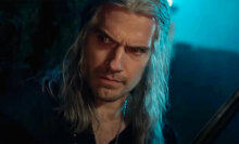 A man with long hair and a sword stands in the darkness.