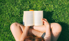 A book open in the lap of a woman sitting in bright green grass. 