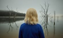 Seen from the back, a young girl with blonde hair wears a blue tracksuit and looks toward a lake. 