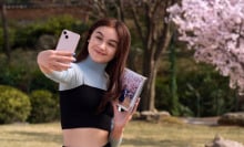 A teenage girl poses for a selfie in front of a cherry blossom tree. 
