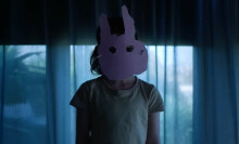 A screenshot of a young girl wearing a poorly cut pink paper rabbit mask, taken from the "Run Rabbit Run" trailer.