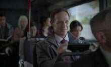 A man smiles while playing a Nintendo Switch on a bus full of people.