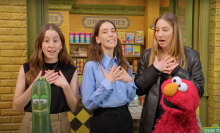 Three women stand on a sidewalk with a red puppet and a zucchini puppet in the foreground.