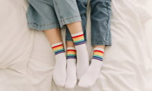 The legs of a lesbian couple both wearing socks with the Pride flag colours, their legs are entwined in an embrace. 