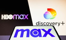 Max logo is seen on a smartphone in front of the HBO Max and Discovery+ logos on a PC screen. 
