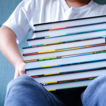 A child sits on a light blue couch holding a large stack of books. 
