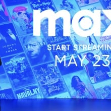 A man in a suit speaks in front of a blue screen covered in TV posters and the words "Max, start streaming May 23."