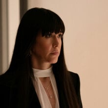 A smartly dressed woman with dark hair stands in a room looking serious.