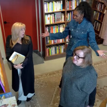 Two women stand in a bookstore, one looking at the shelves while another looks surprised by a third woman appearing behind them.
