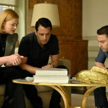Two men and a woman wearing smart clothes sit around a table.