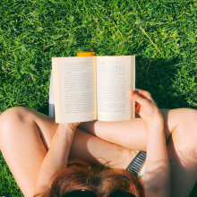 A book open in the lap of a woman sitting in bright green grass. 