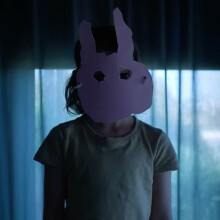 A screenshot of a young girl wearing a poorly cut pink paper rabbit mask, taken from the "Run Rabbit Run" trailer.