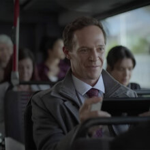 A man smiles while playing a Nintendo Switch on a bus full of people.
