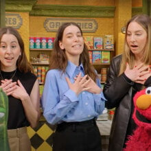 Three women stand on a sidewalk with a red puppet and a zucchini puppet in the foreground.