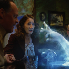 A group of adults sit around a table, with a ghost's hand coming out of its center. 