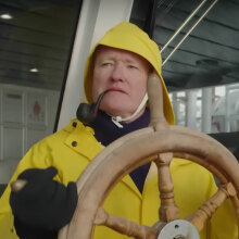 A man stands behind the wheel of a boat wearing a yellow rain jacket with the hood up. He has a pipe in his mouth.