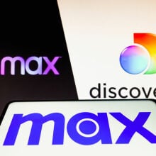 Max logo is seen on a smartphone in front of the HBO Max and Discovery+ logos on a PC screen. 