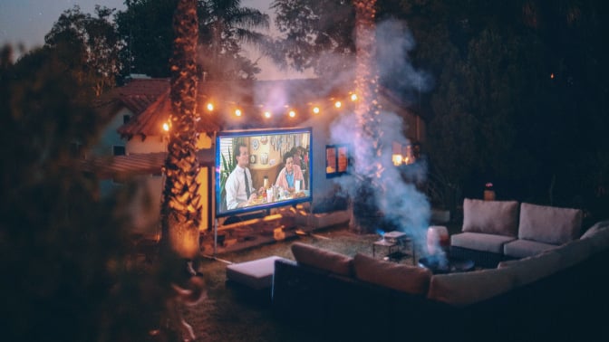 Screen on side of house surrounded by fairy lights showing video and facing set of couches around a fire