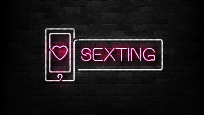 A neon sign with pink text which reads "sexting" on a black brick wall.