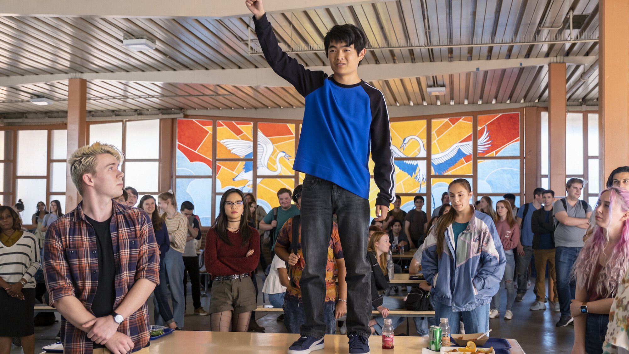 A teenager wearing a blue shirt stands atop a table in a high school cafeteria.