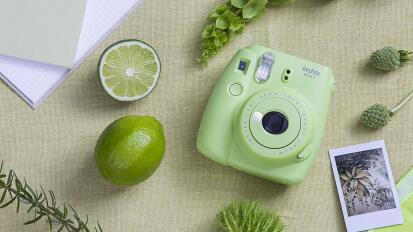 a green Instax camera on a table, surrounding by other green items like a lime and sprigs of greenery