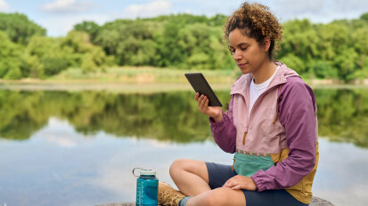 person reading on a Kindle Paperwhite on a dock by a lake