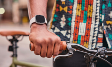 a close-up of a fitbit sense 2 on a person's wrist as they hold a bike's handlebar