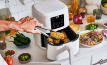 white air fryer with, hand removing corn with metal tongs. Air fryer is in a kitchen counter with produce.