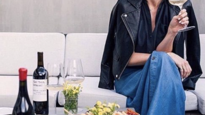 a woman in a leather jacket holding a glass of wine on her sofa, with a bottle of wine on the table in front of her