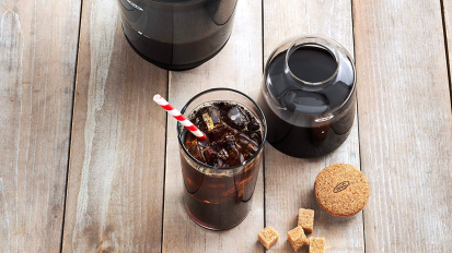 a top-down view of the OXO Brew Compact Cold Brew Coffee Maker, a glass of iced coffee with a striped straw, and brown sugar cubes on a rustic wood table