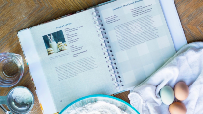 a createmycookbook recipe book surrounded by a bowl of flour, measuring cups, and three eggs sitting on a hand towel