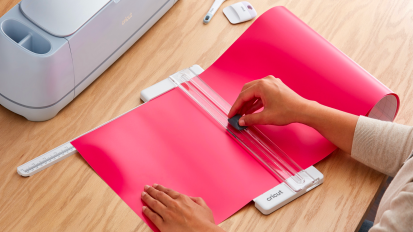a close-up of a woman using a paper cutter to cut a piece of cardstock next to a cricut machine on a wood table