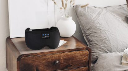a bluetooth-enabled sleep mask on a wooden dresser next to a bed with beige linens and a white vase