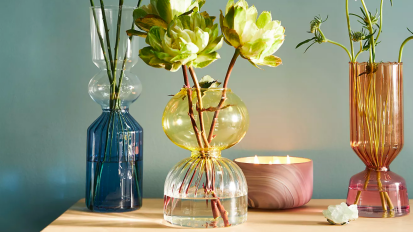 three anthropologie calle vases filled with flowers on a wooden dresser next to a lit three-wick candle