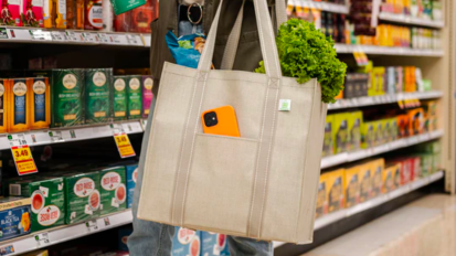 a close-up of a woman in a grocery store holding a beige reusable grocery bag filled with lettuce