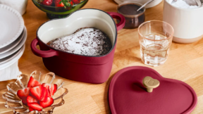 the Beautiful 2QT Cast Enamel Heart Dutch Oven filled with a chocolate cake next to ingredients for chocolate-covered strawberries on a wooden countertop