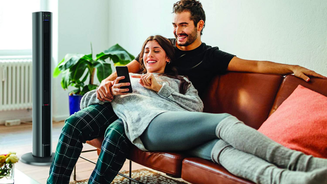 a couple sitting on a tan couch in front of a Lasko FH500